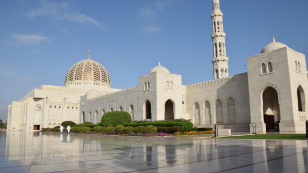 Weiße Moschee mit Kuppel und Minarett vor blauem Himmel, davor spiegelnder Platz und Blumenbeete.