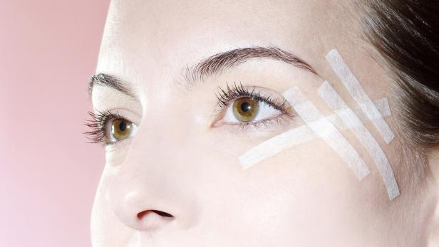 Das Gesicht einer Frau mit Pflastern auf der Schläfe vor einem rosa Hintergrund.