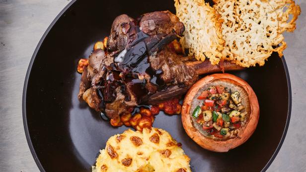 Ein Teller mit Lammkeule, gefüllter Zwiebel, Kartoffelgratin und Käse-Crackern.