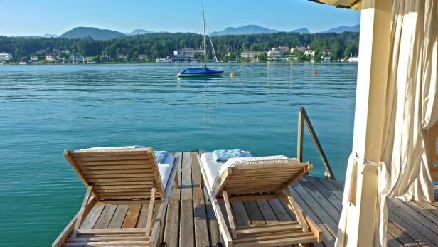 Zwei Liegestühle stehen auf einem Holzsteg mit Blick auf einen See und bewaldete Hügel.