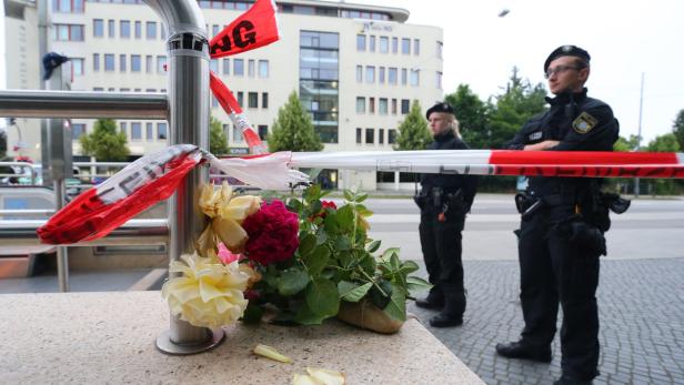 Blumen liegen an einem Tatort, der mit rot-weißem Band abgesperrt ist; zwei Polizisten stehen Wache.
