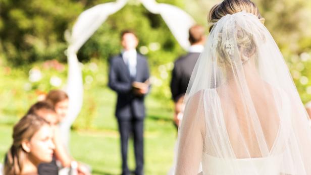 Eine Braut mit Schleier steht im Freien vor einem Traubogen, während Gäste und ein Redner warten.