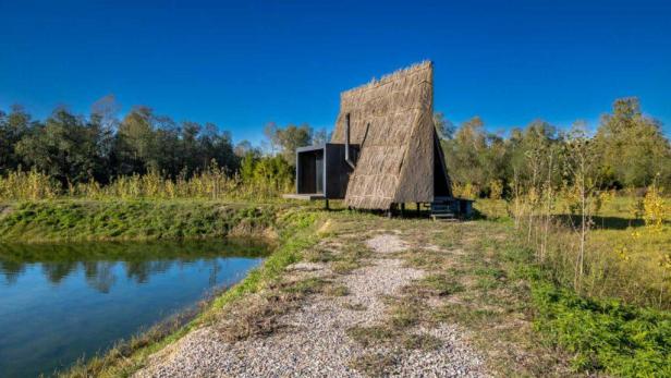 Ein strohgedecktes Haus mit einem modernen Anbau steht an einem kleinen See.