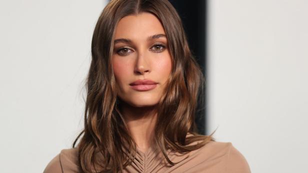 Porträt von Hailey Bieber mit braunem, gewelltem Haar und einem beigen Oberteil.