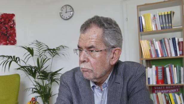 Ein älterer Mann mit Brille sitzt in einem Raum mit Bücherregal und Zimmerpflanze.