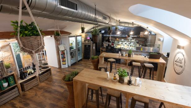 Innenansicht eines „Little Italian Deli“-Geschäfts mit Tischen und einer Theke.