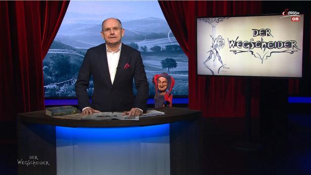 Ein Mann steht an einem Tisch vor einem Bildschirm mit dem Titel „Der Wegscheider“.