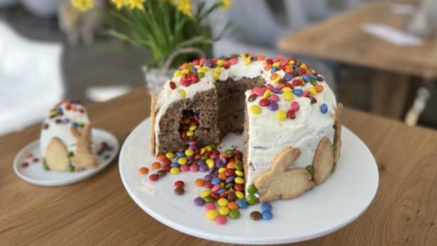 Ein angeschnittener Osterkuchen mit bunten Schokolinsen und Hasenkeksen steht auf einem Tisch.