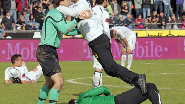 Zwei Fußballspieler umarmen sich auf dem Spielfeld, während andere Spieler im Hintergrund zu sehen sind.