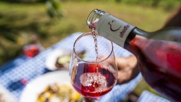 Uhudler-Wein wird aus einer Flasche in ein Weinglas gegossen, während im Hintergrund eine karierte Tischdecke und Speisen zu sehen sind.