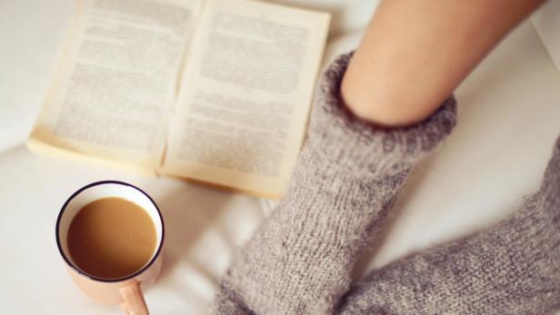 Ein Paar Wollsocken, eine Tasse Kaffee und ein offenes Buch auf einem Sofa.