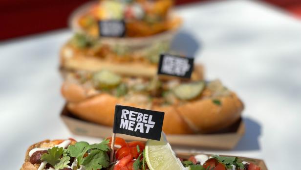 Drei Hotdogs von „Rebel Meat“ mit verschiedenen Toppings werden auf einem Tisch präsentiert.
