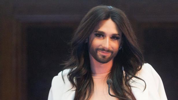 Conchita Wurst lächelt in einem weißen Blazer in die Kamera.