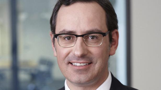 Ein lächelnder Mann mit Brille und dunklem Anzug vor einem unscharfen Hintergrund.