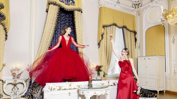 Zwei Frauen in roten Kleidern feiern in einem opulenten Schlafzimmer mit einem Glas Sekt.