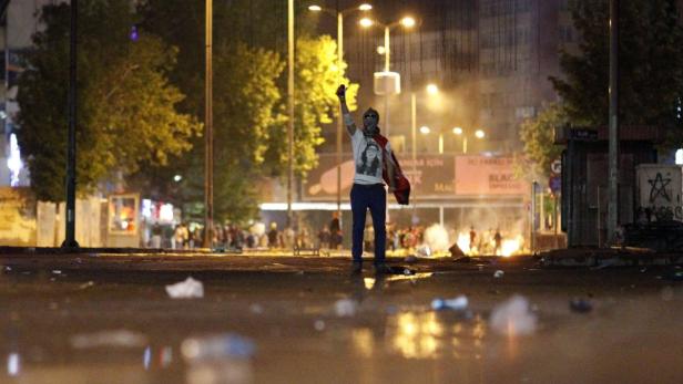 Ein Demonstrant steht nachts auf einer Straße mit erhobener Faust, im Hintergrund brennen Barrikaden.