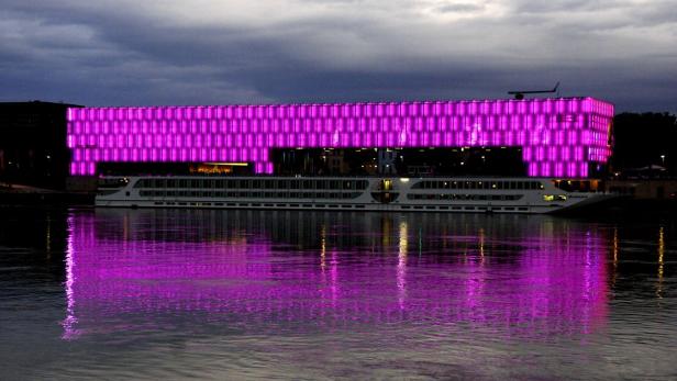 Ein Flusskreuzfahrtschiff liegt vor einem Gebäude, das mit pinkfarbenen Lichtern beleuchtet ist.
