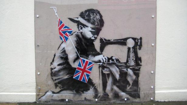 Ein Graffito zeigt ein Kind, das an einer Nähmaschine mit Union-Jack-Flaggen arbeitet.