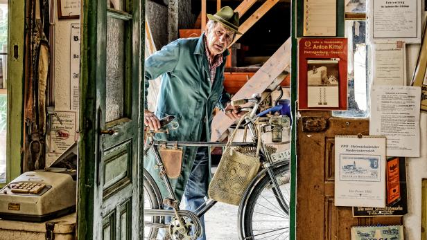 Ein älterer Mann mit Hut schiebt sein Fahrrad durch eine offene Tür in einen Laden.