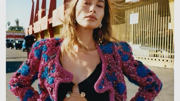 Hailey Bieber posiert in einer bunten Jacke und einem schwarzen Top vor einem Gebäude.