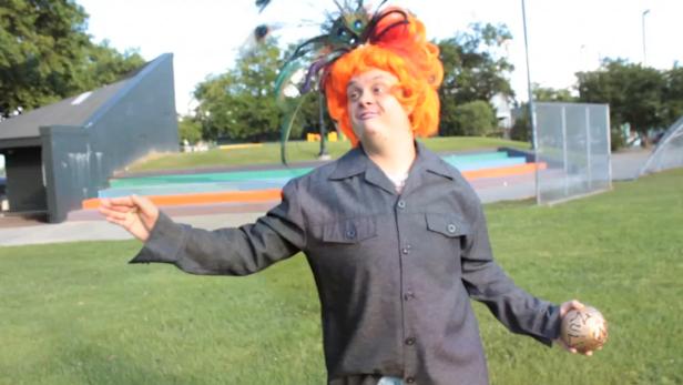 Eine Person mit orangefarbener Perücke und Federschmuck posiert mit einer goldenen Kugel in einem Park.