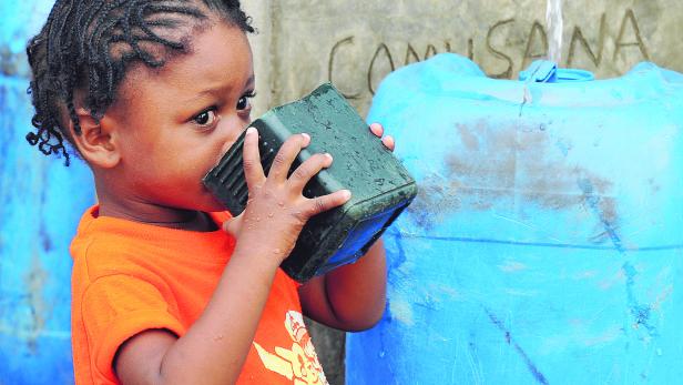 Ein kleines Mädchen trinkt Wasser aus einem Becher vor einem Hilfswerk-Brunnen.