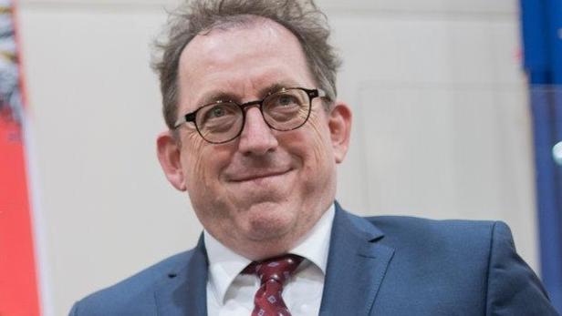 Ein älterer Mann hält eine rote Mappe des Presseclub Concordia und einen Blumenstrauß, flankiert von zwei Männern.