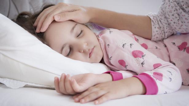 Ein schlafendes Kind liegt im Bett, die Mutter greift ihm auf die Stirn.