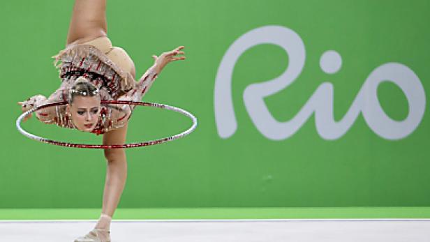 Eine rhythmische Sportgymnastin führt eine Übung mit einem Reifen vor dem Hintergrund des „Rio“-Logos auf.
