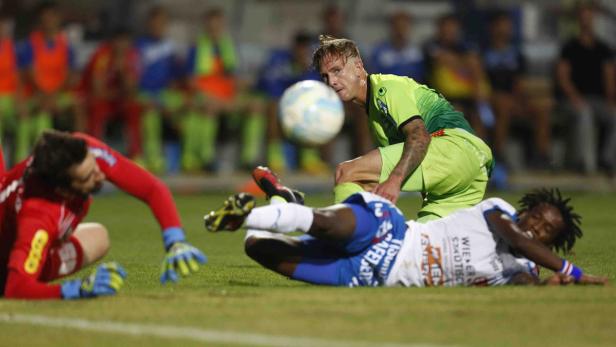 Ein Fußballspieler in Grün versucht, den Ball vor einem am Boden liegenden Gegenspieler zu erreichen.