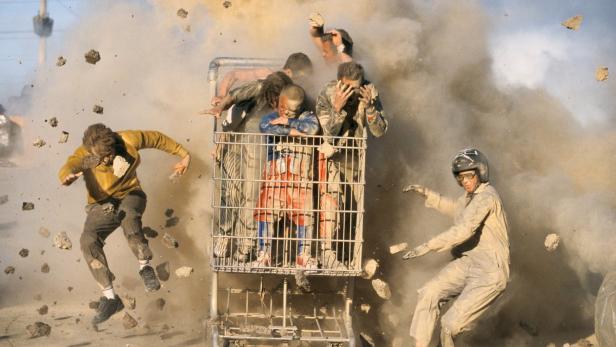 Mehrere Personen in einem Einkaufswagen inmitten einer Staubwolke und umherfliegender Trümmer.