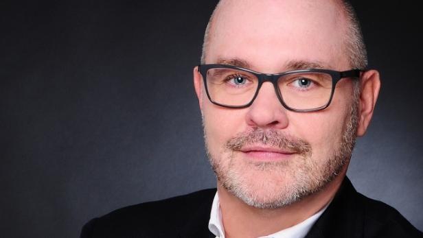 Ein Mann mit Brille und Anzug vor einem dunklen Hintergrund.