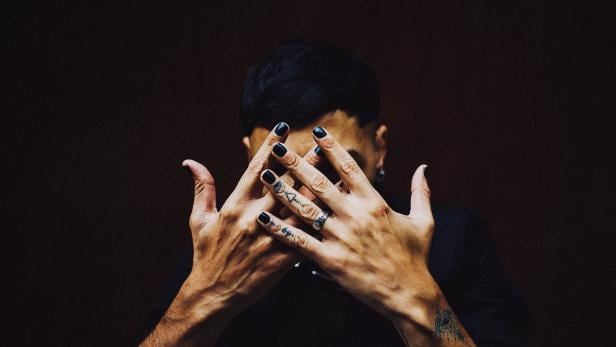 Eine Person mit Tattoos und schwarz lackierten Nägeln verdeckt ihr Gesicht mit den Händen.