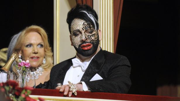 Ein Mann mit aufwendigem Make-up und eine Frau mit Schmuck sitzen nebeneinander.