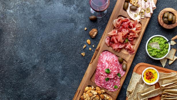 Eine Holzplatte mit Wurst, Käse, Nüssen, Oliven, Dips und zwei Gläsern Rotwein.