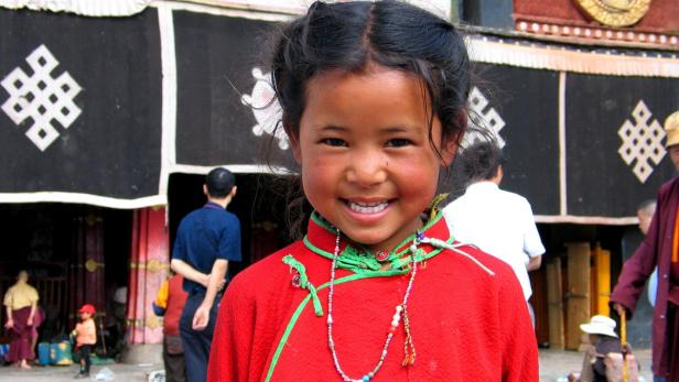 Ein lächelndes Mädchen in traditioneller Kleidung vor einem Gebäude in Tibet.