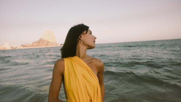 Eine Frau in einem gelben Kleid steht am Strand, im Hintergrund ein Felsen im Meer.