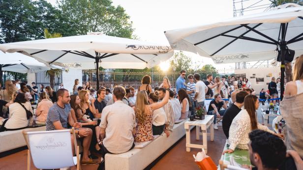 Viele Menschen sitzen und stehen unter Sonnenschirmen bei einer Veranstaltung im Freien.