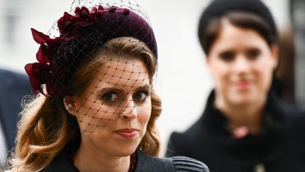 Prinzessin Beatrice mit langem Haar trägt einen weinroten Hut mit Schleier und Blumen, im Hintergrund steht Prinzessin Eugenie unscharf.