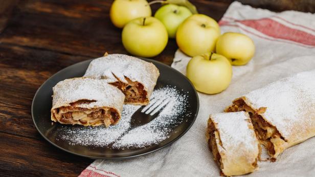 Apfelstrudel mit Puderzucker auf einem Teller mit Äpfeln im Hintergrund.