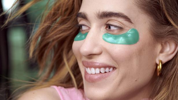 Eine Frau lächelt mit grünen Augenpads und goldenen Ohrringen.