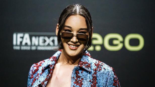 Model Alex Mariah Peter mit Sonnenbrille trägt ein blau-rot gemustertes Hemd vor einem dunklen Hintergrund.