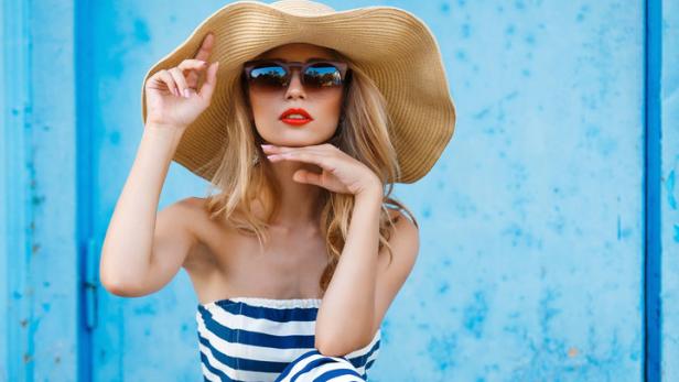 Eine Frau mit Hut und Sonnenbrille posiert in einem blau-weiß gestreiften Kleid vor einer blauen Wand.