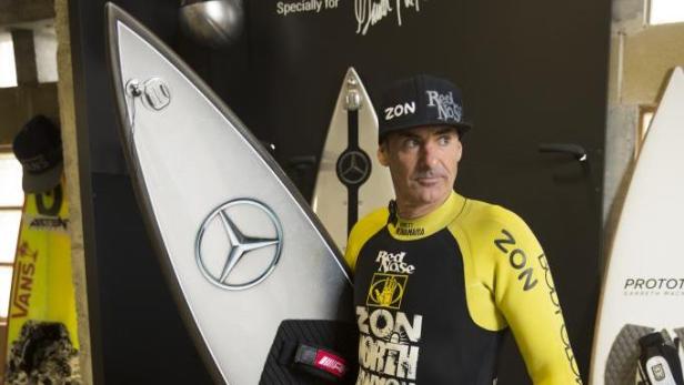 Ein Mann mit Kappe und Neoprenanzug steht neben einem Surfbrett mit Mercedes-Logo.