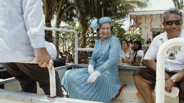 Königin Elisabeth II. sitzt in einem offenen Fahrzeug, umgeben von Menschen und Palmen.