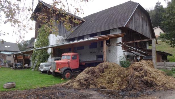 Ein roter Lastwagen steht vor einem alten Bauernhaus mit einem großen Misthaufen.