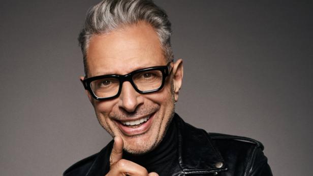 Jeff Goldblum lächelt in schwarzer Lederjacke und Brille, den Finger an die Wange gelegt.