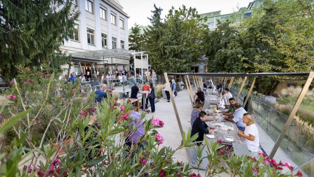 Menschen sitzen an Tischen im Freien, umgeben von blühenden Büschen.