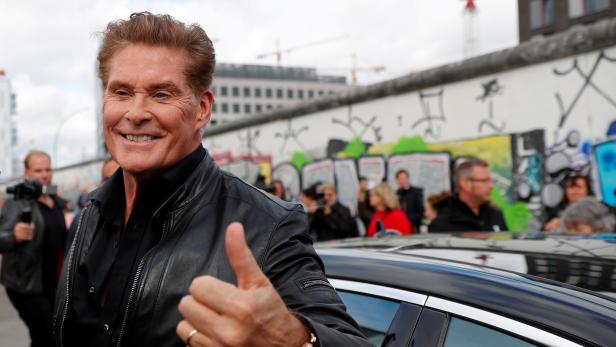 David Hasselhoff lächelt und zeigt den Daumen hoch vor einem Stück der Berliner Mauer.