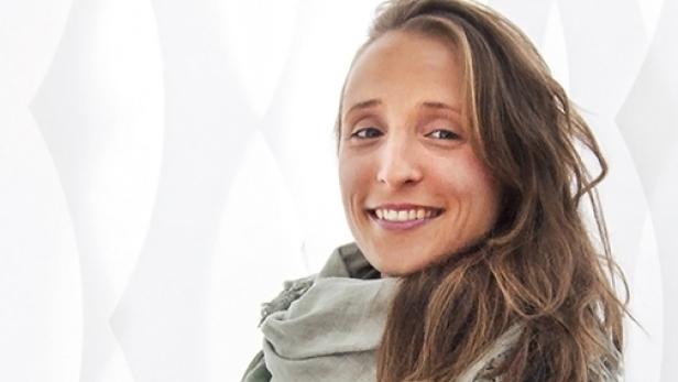 Eine lächelnde Frau mit langen, braunen Haaren und einem grünen Schal vor einem weißen Hintergrund.